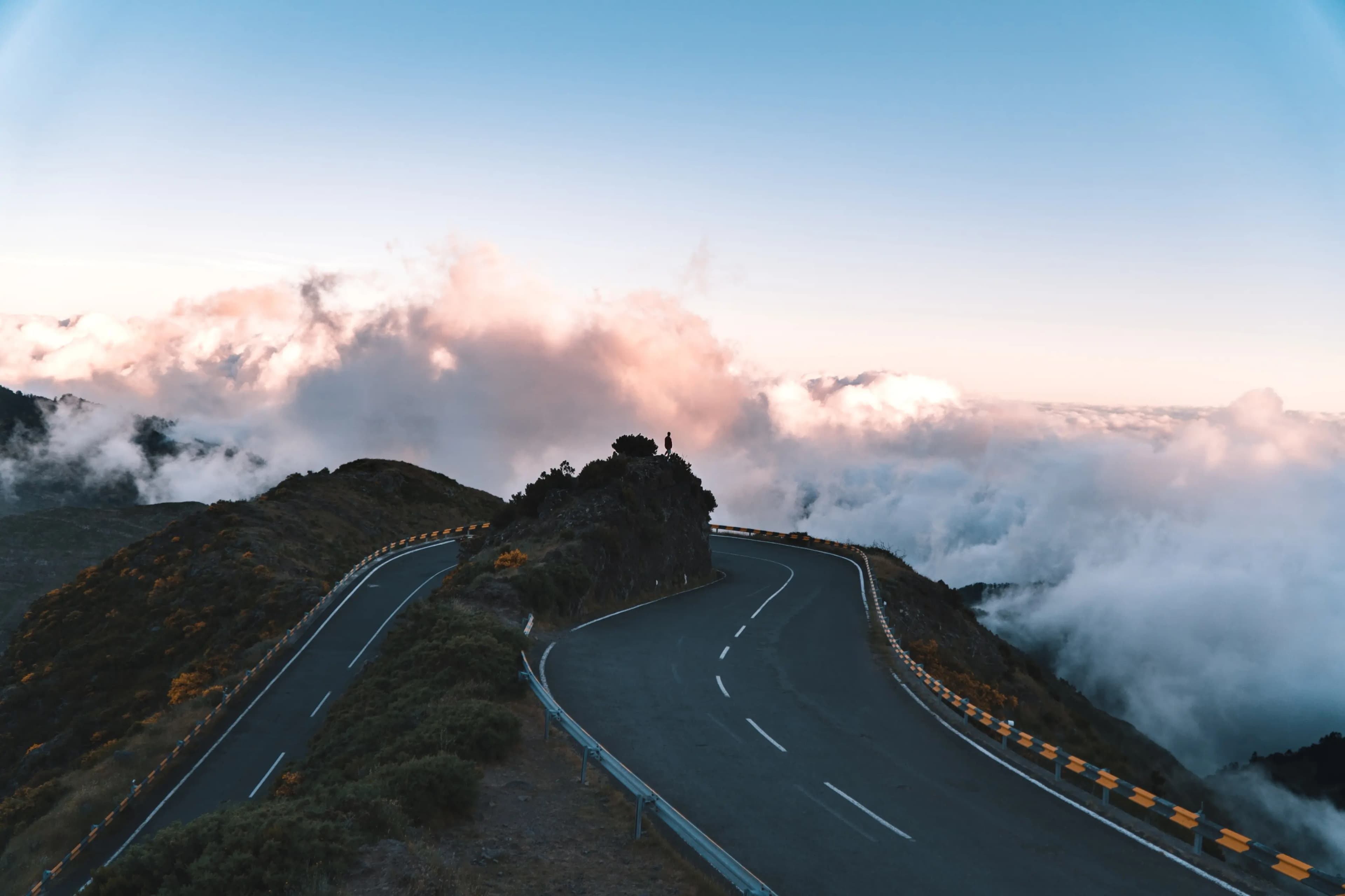 Person genießt die Aussicht auf eine kurvige Bergstraße über den Wolken beim Sonnenuntergang – finde solche atemberaubenden Landschaften und Traumrouten mit unserer Motorradnavigation
